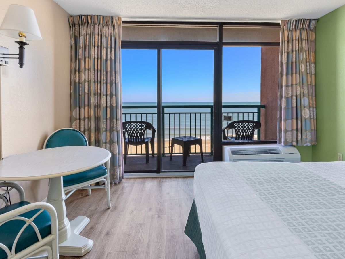 A hotel room with a bed, small table with chairs, and a view of the ocean from a balcony with patio furniture, curtains drawn to each side.