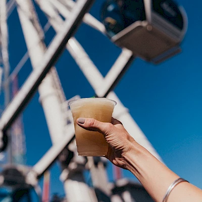 A hand holding a drink in front of a Ferris wheel on a bright, clear day, with the blue sky in the background.