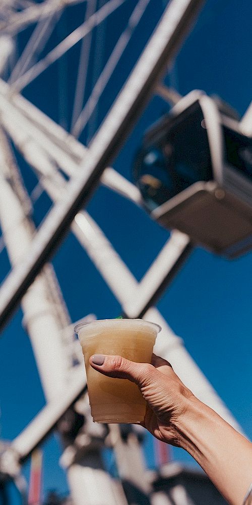 A hand holding a drink in front of a Ferris wheel on a bright, clear day, with the blue sky in the background.