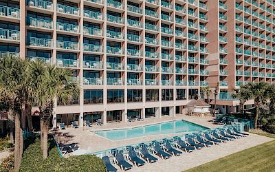 The image shows a multi-story hotel building with balconies overlooking a swimming pool area lined with lounge chairs and bordered by palm trees.
