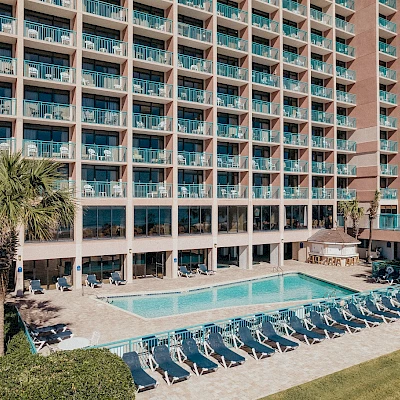 The image shows a multi-story hotel building with balconies overlooking a swimming pool area lined with lounge chairs and bordered by palm trees.