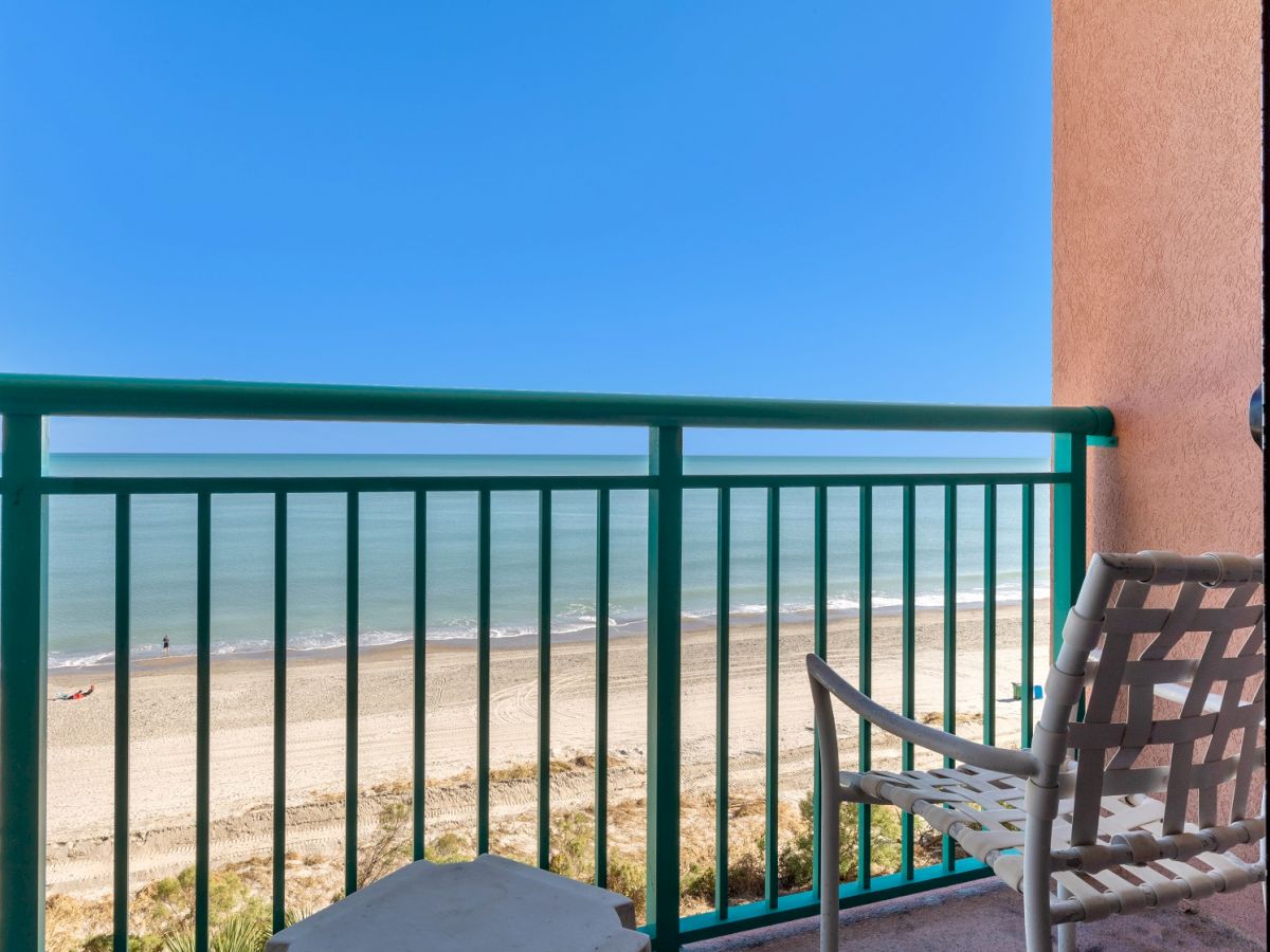 A balcony overlooking a sandy beach with a blue ocean. There is a chair and a small table on the balcony, framed by green railing.