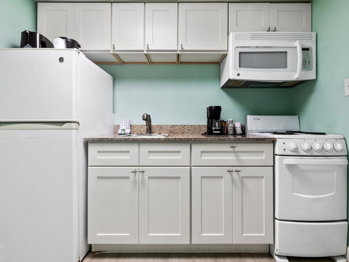 A compact kitchen featuring a white refrigerator, microwave, stove, sink, coffee maker, and cabinetry on a mint green wall.