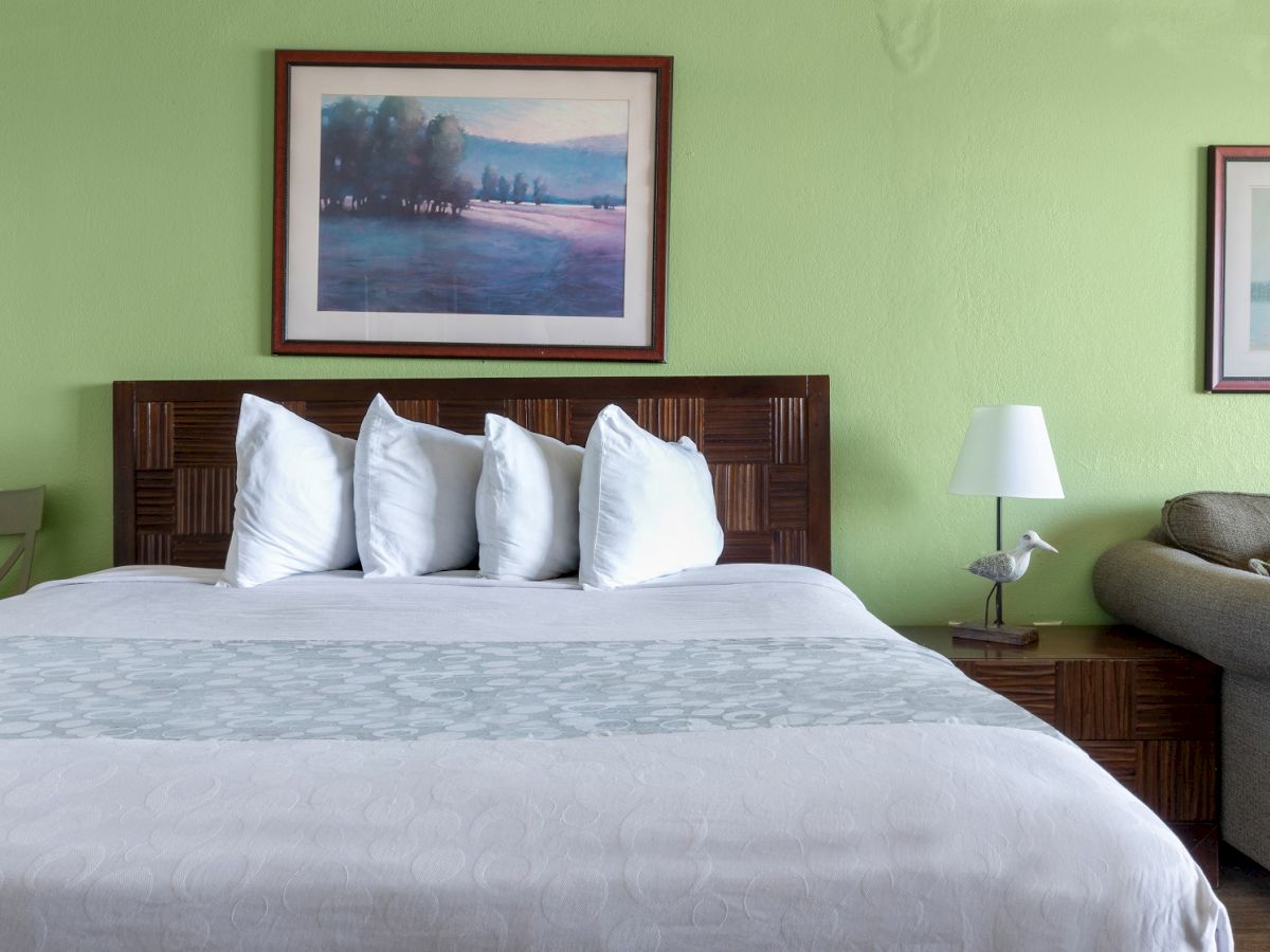 A neatly made bed with four pillows, framed artwork on green walls, a lamp on a nightstand, and a green couch next to the bed ends the sentence.