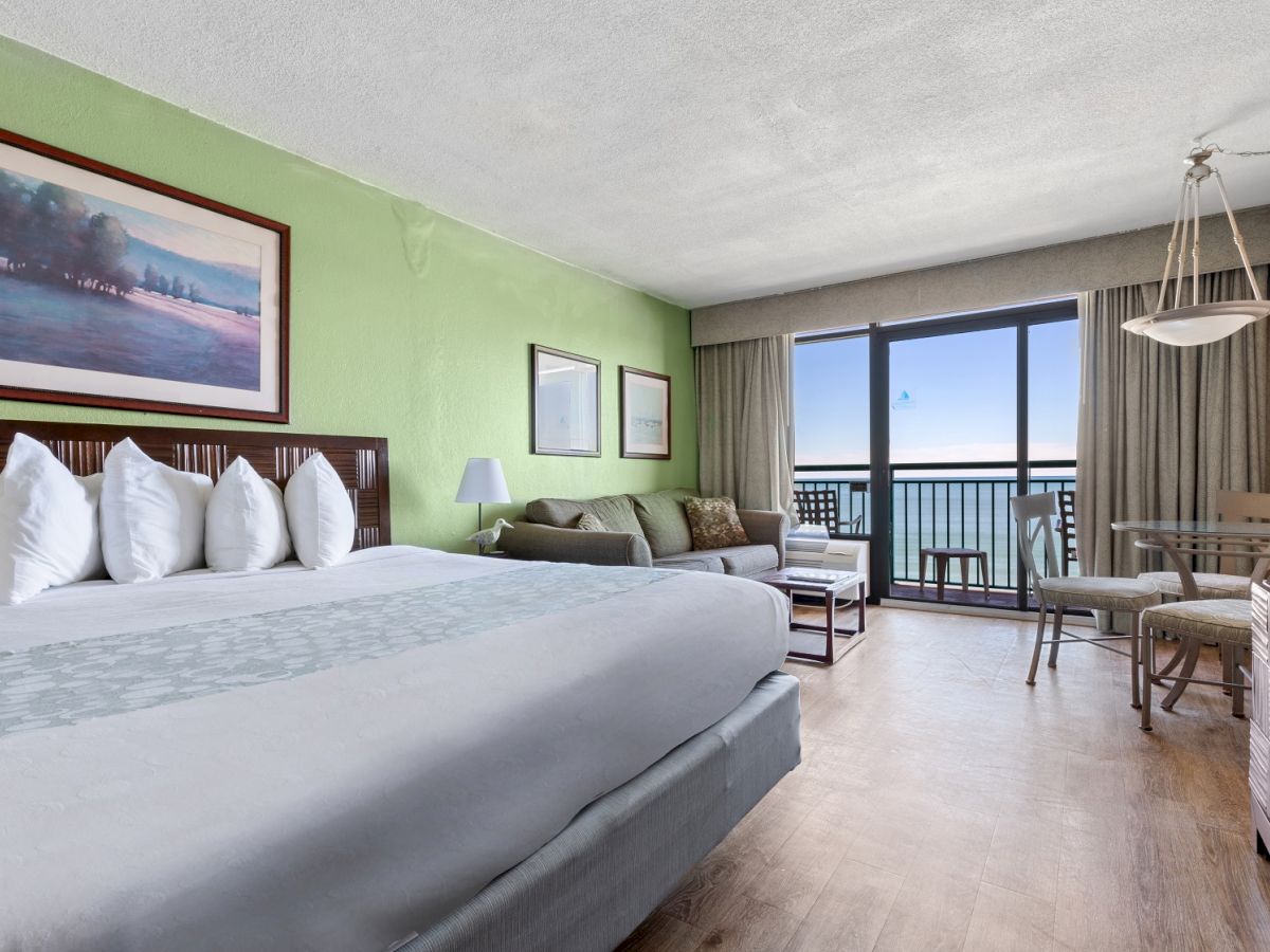 The image shows a spacious hotel room with a large bed, a sitting area, a balcony with a sea view, and modern furniture and decor.