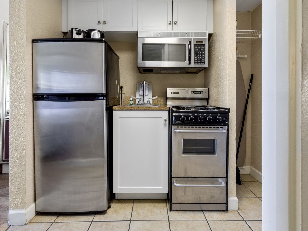 A compact kitchen with a stainless steel fridge, microwave, stove, and white cabinetry. A kettle and coffee maker sit on the counter.