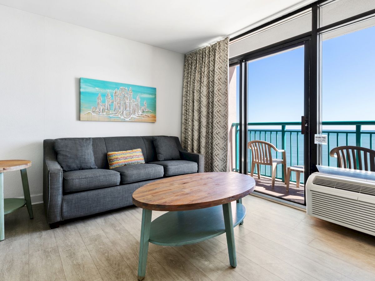 A living room with modern furniture, seaside artwork, a coffee table, and an ocean-view balcony with chairs is shown in the image.
