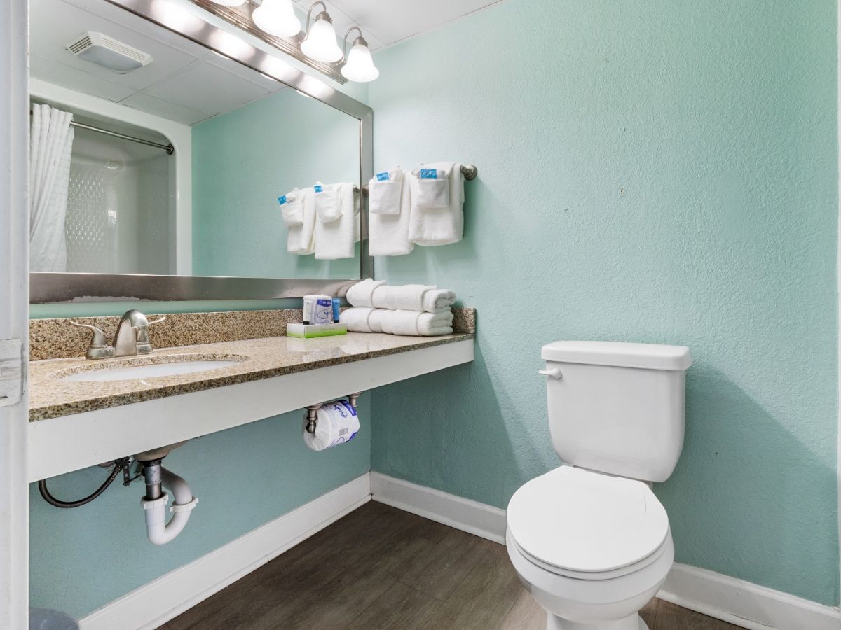 A bathroom with a granite countertop, sink, mirror with lights, toilet, and neatly folded towels on a mint green wall.