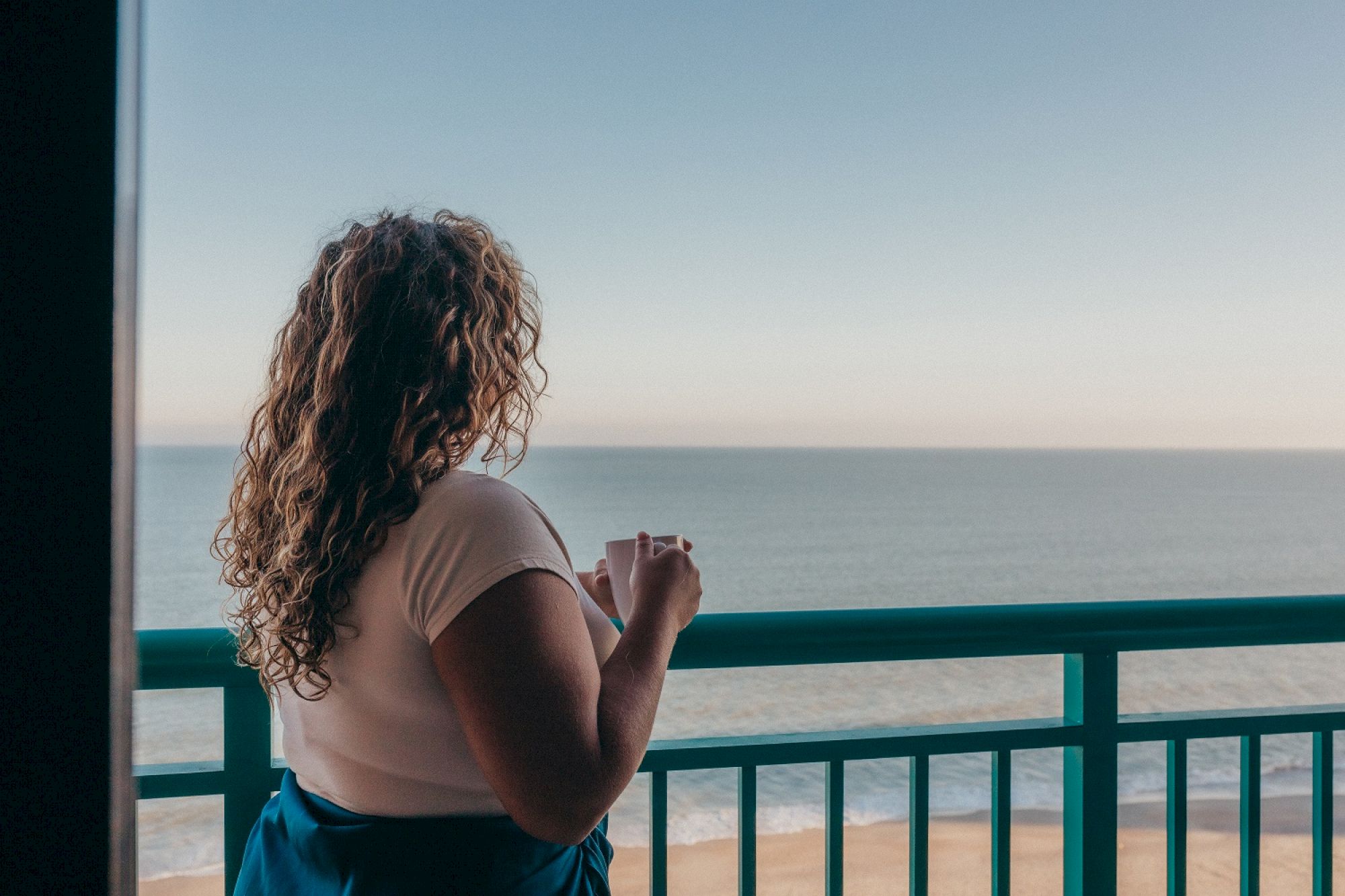 A person with curly hair stands on a balcony, gazing out at the ocean under a clear sky.