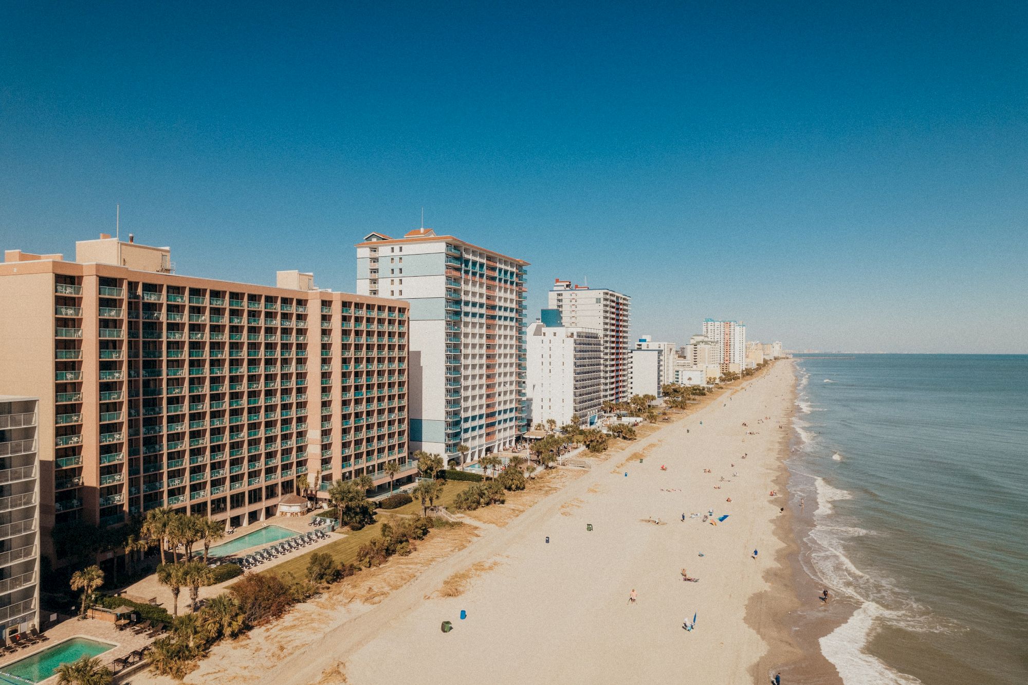 A coastline with tall buildings lining a sandy beach, with a calm ocean and clear blue sky in the background.