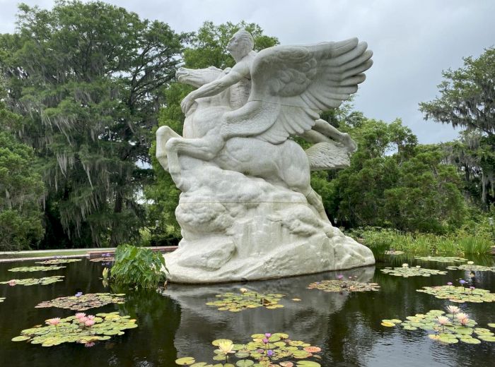 A statue depicting a winged horse and rider, surrounded by lily pads, is set in the middle of a water feature with trees in the background.