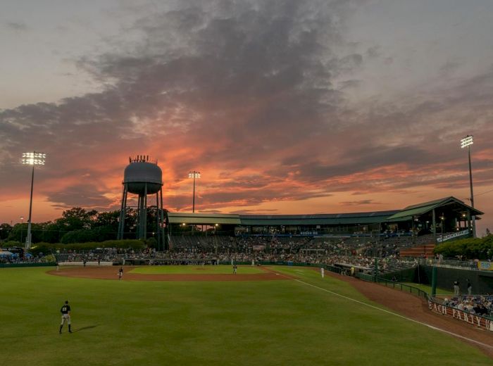 A baseball field at sunset with players on the field and a stadium in the background, along with a water tower lit by stadium lights.