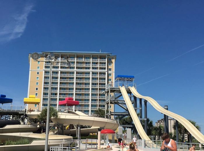 The image shows a water park with slides, a pool, and a high-rise building in the background. People are sunbathing and enjoying the sunny day.