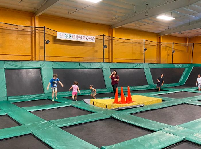 Children and adults are enjoying a trampoline park with green and black trampolines and orange cones. The room has bright yellow walls.