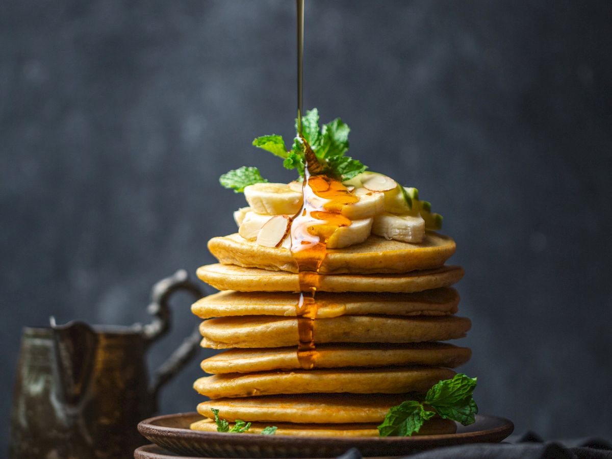 A stack of pancakes topped with banana slices and mint leaves, being drizzled with syrup from a metal container.
