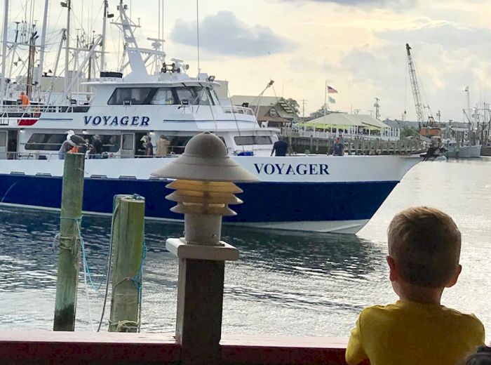 A child in a yellow shirt watches a boat named VOYAGER pass by at a marina. There are structures and other boats in the background.