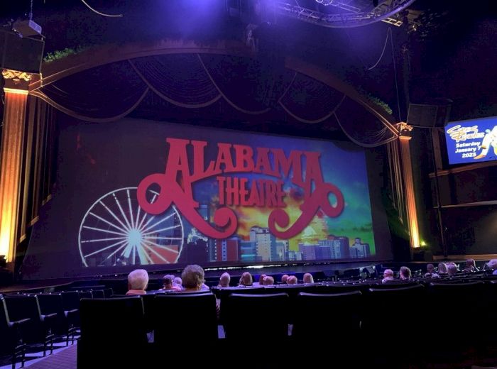 An audience sits in a theater, looking at a stage with a large sign reading "Alabama Theatre." The stage background features a Ferris wheel and cityscape.