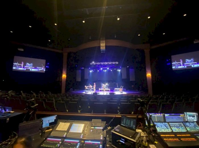 The image shows a theater or concert hall with a stage lit in blue and a control panel in the foreground, likely used for sound and lighting.