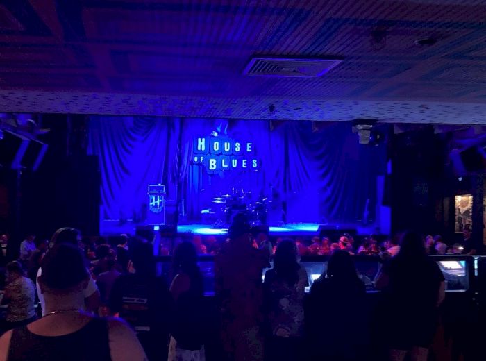 The image shows an indoor concert venue with a stage set up and a dark crowd gathered. A "House of Blues" sign is illuminated in the background.