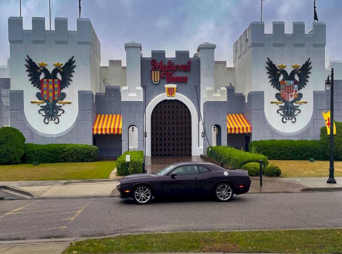 A black car is parked in front of a castle-themed building with "Medieval Times" signage and flags, surrounded by greenery and a cloudy sky.