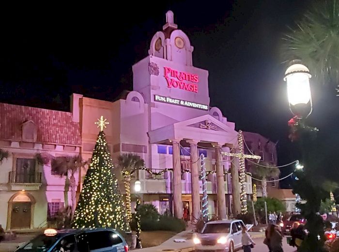 A brightly lit building with a sign that reads "Pirates Voyage Dinner & Show," adorned with holiday decorations, cars, and a Christmas tree at night.