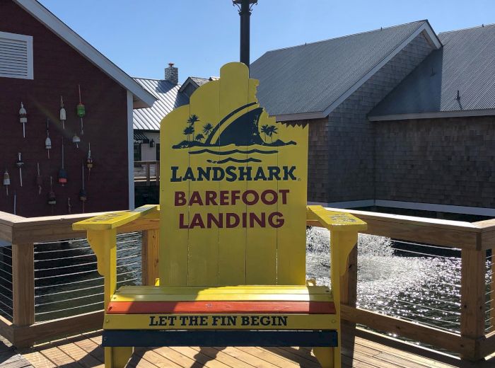 The image shows a large, yellow chair with the text "LANDSHARK BAREFOOT LANDING" and hashtags "LET THE FIN BEGIN" and "#LANDSHARKBARANDGRILL".