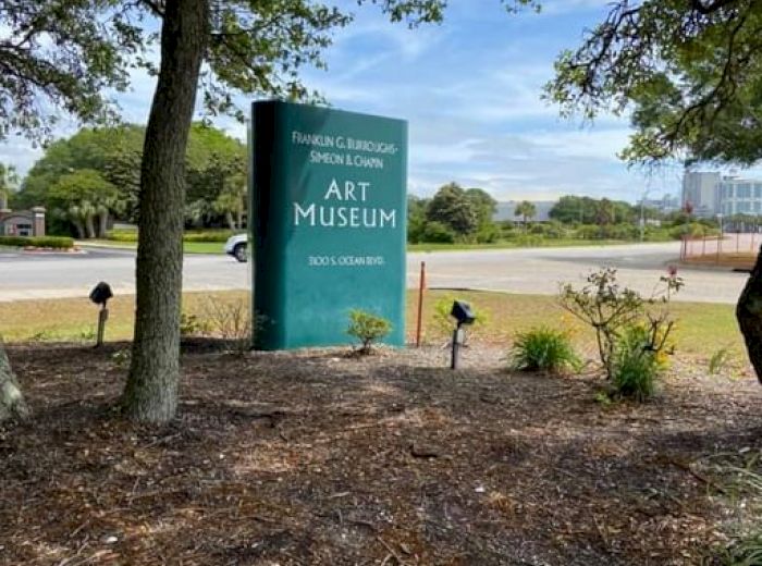 The image shows a green sign for the Rockport Center for the Arts Museum, located outdoors among trees and plants, with a parking lot in the background.