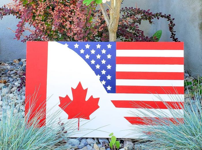 The image shows a sign combining the American and Canadian flags, placed in a garden with decorative plants and rocks in the background.