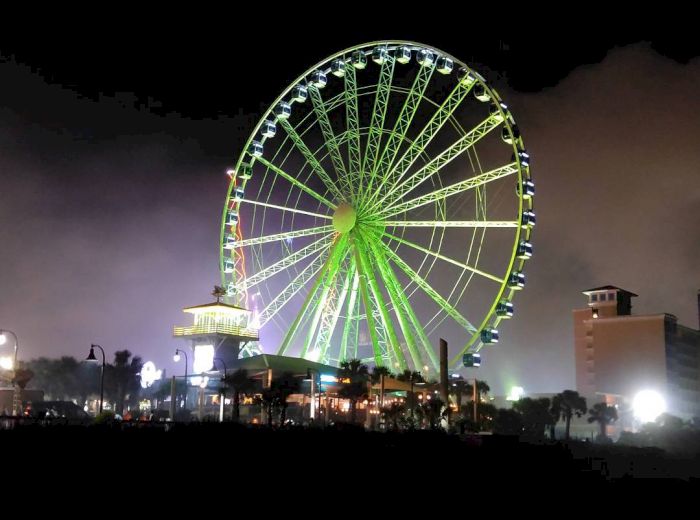 A brightly lit Ferris wheel glowing green at night with surrounding buildings and lights in the background.