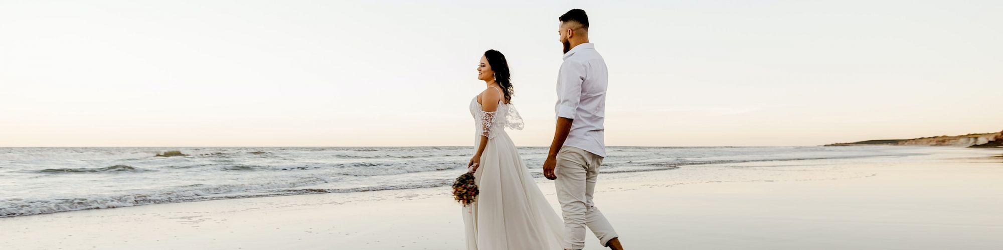 A couple is walking barefoot on a beach near the water’s edge during sunset, with the woman in a white dress and the man in light-colored clothing.