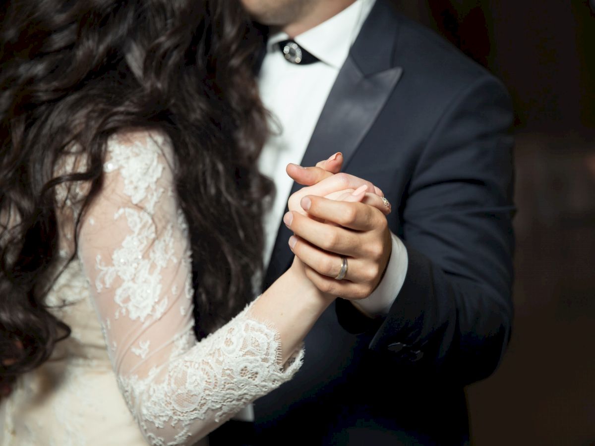 A couple dressed in formal attire, with the woman in a lace dress and the man in a suit, hold hands while dancing.