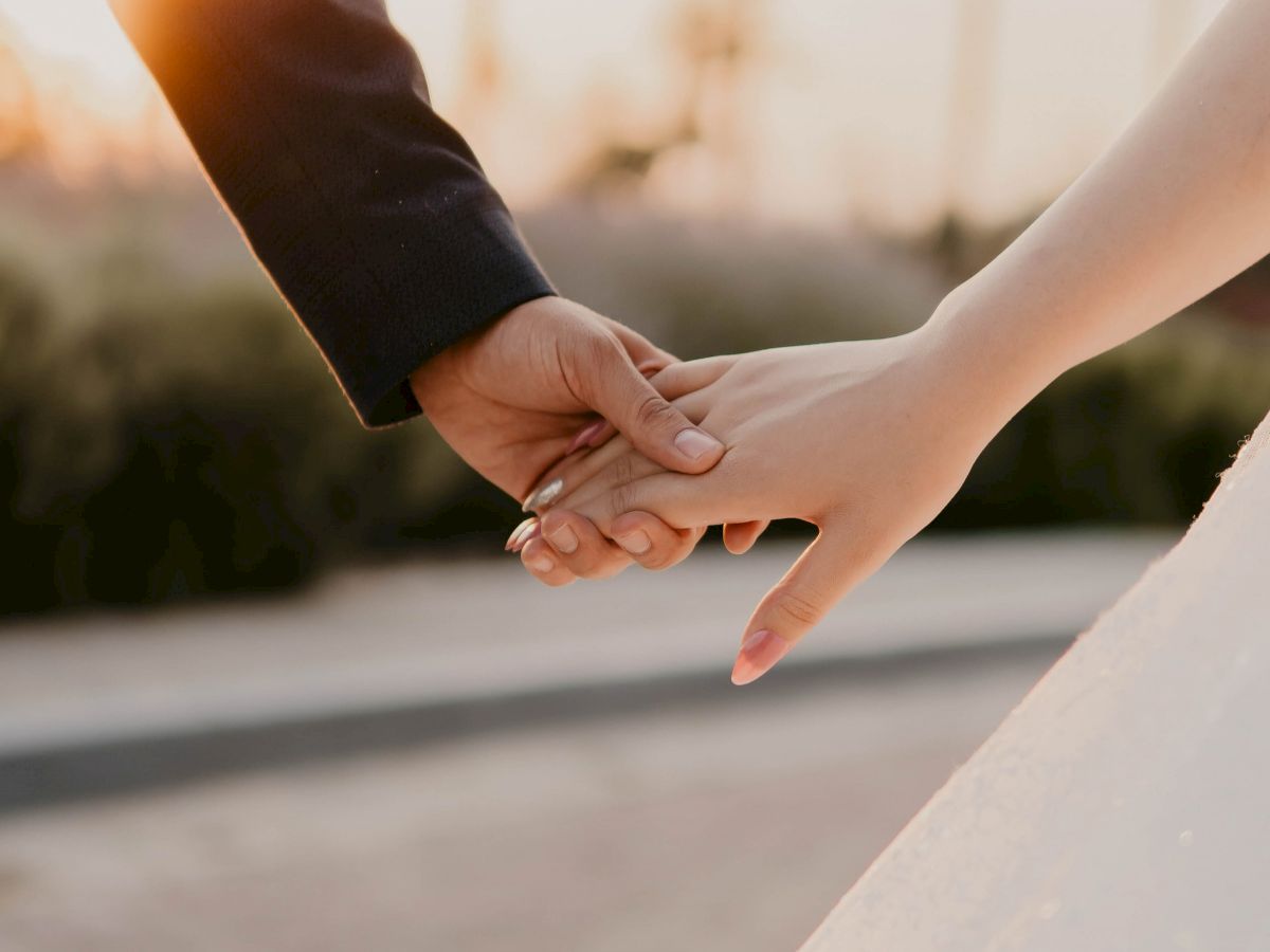 A couple is holding hands outdoors during sunset, one person in dark attire, and the other in a white dress.