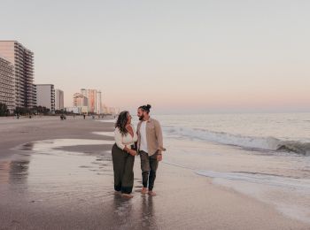 A couple is standing on the beach near the shoreline with high-rise buildings in the background at sunset, holding hands and looking at each other.