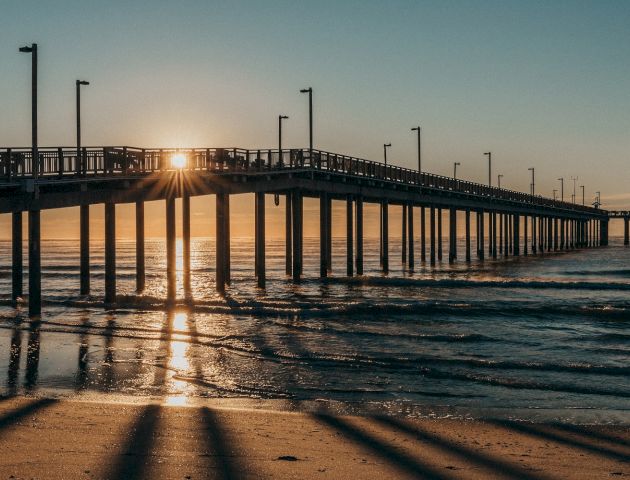 A long pier extends over the ocean with the sun setting behind it, casting long shadows on the sandy beach below, captured during the golden hour.
