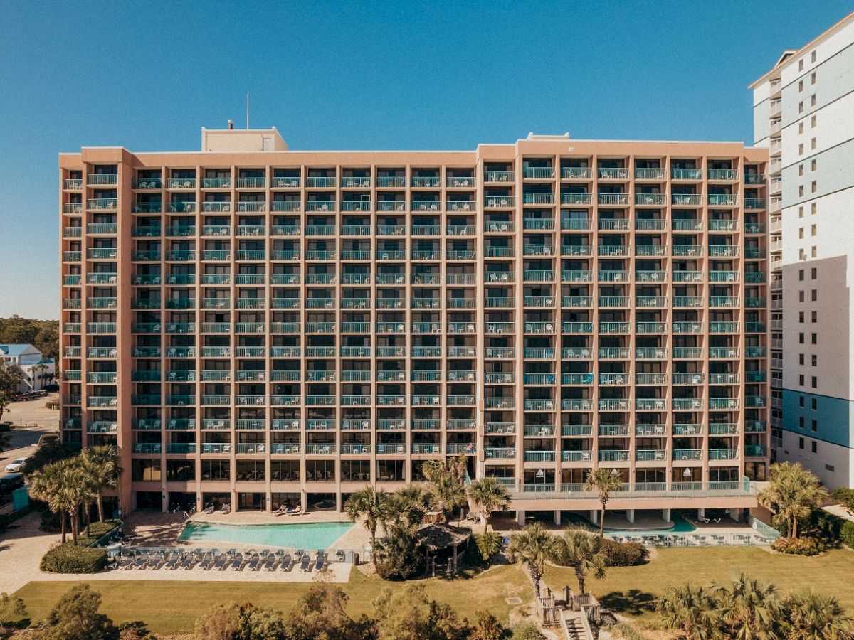 The image shows a large multi-story apartment or hotel building with balconies, a swimming pool area, and palm trees in front.