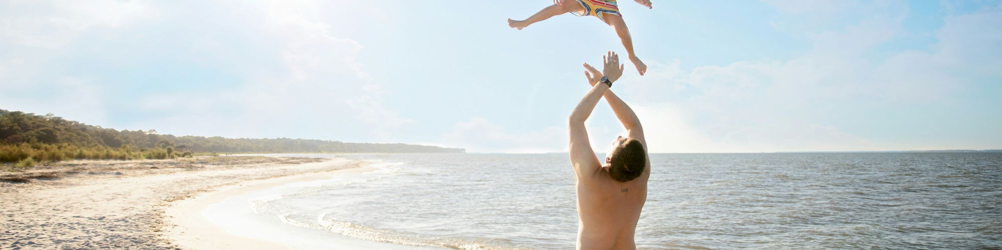 An adult is playing with a child by tossing them into the air on a sunny beach, with the ocean and shoreline in the background.