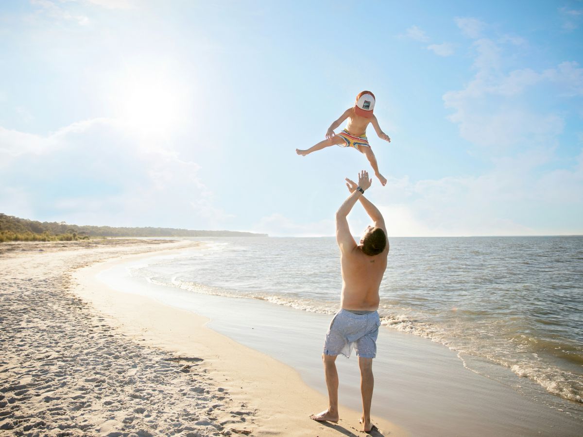 A man is playing with a child on a sunny beach, joyfully lifting them into the air near the shoreline, both appearing happy and carefree.
