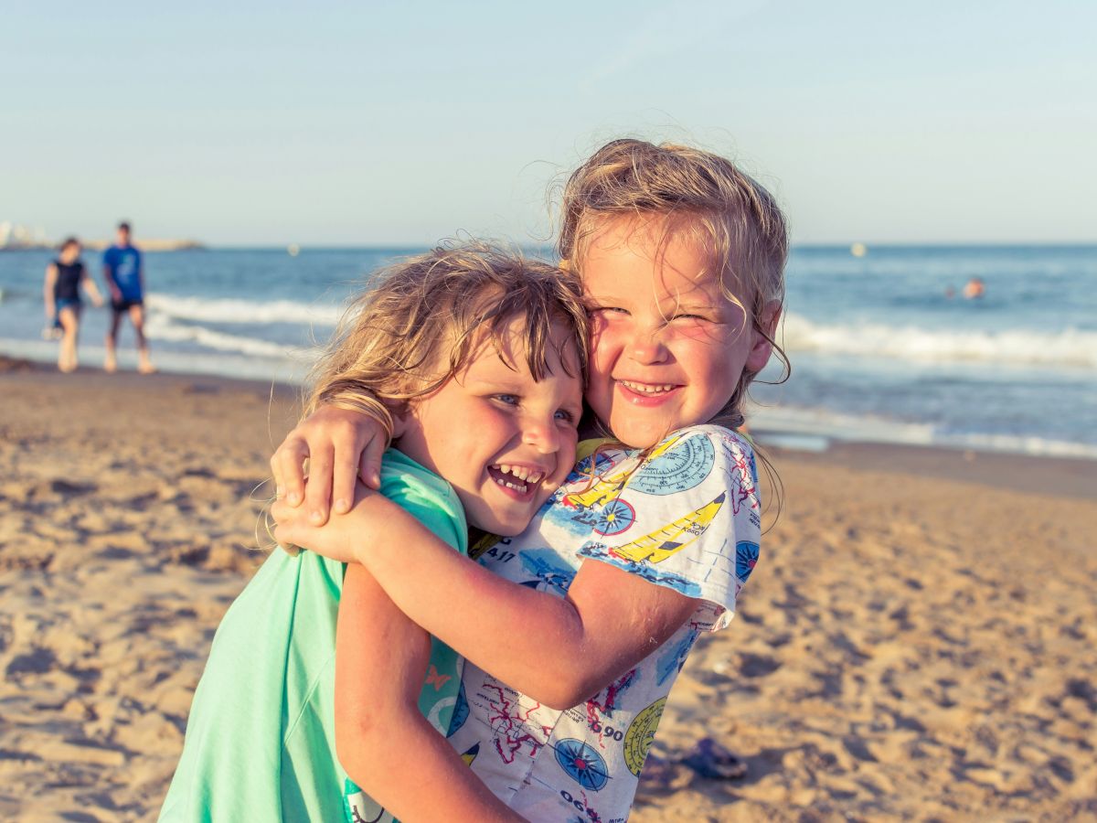 Two children enjoying a warm hug on the beach, with the ocean and a few people in the background on a sunny day.