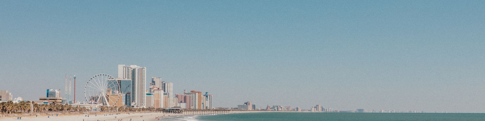 The image shows a beach with a few people walking near the shore, with a city skyline and Ferris wheel in the background.