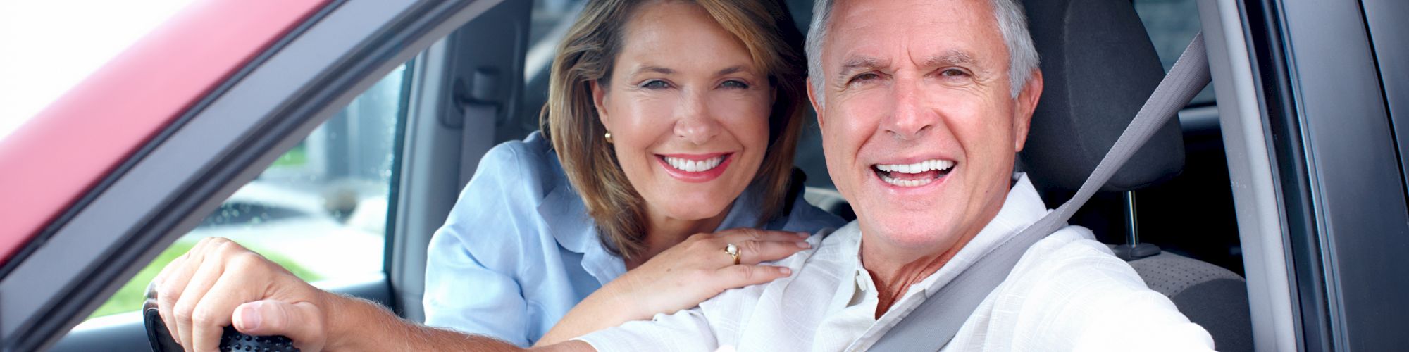 A smiling man and woman are sitting in the front seats of a red car, with the man giving a thumbs-up gesture.