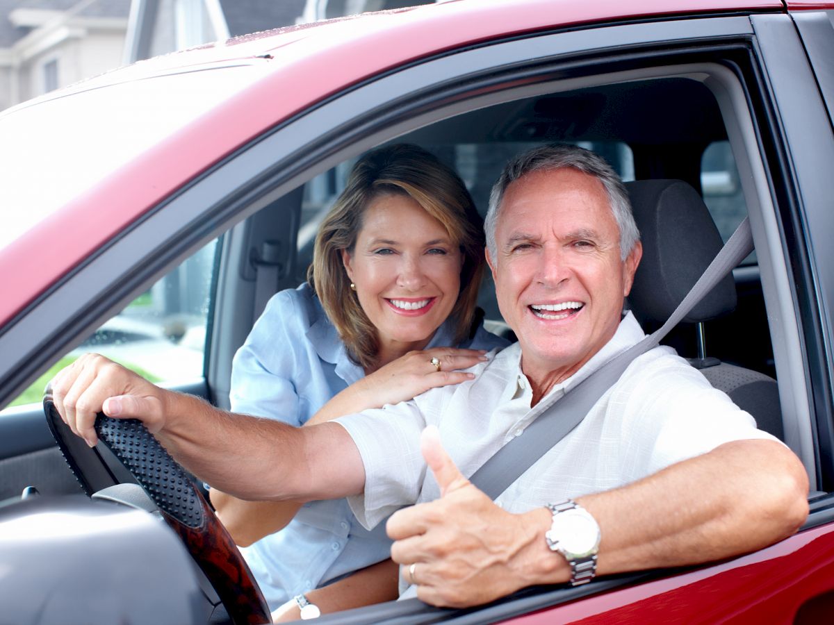 A smiling couple is sitting in a red car, with the man in the driver's seat giving a thumbs-up and the woman leaning in from the passenger side.