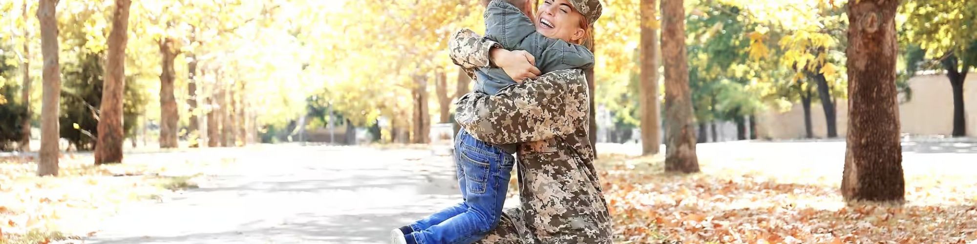 A person in military uniform embraces a child in a tree-lined pathway filled with autumn leaves, both appearing joyful.