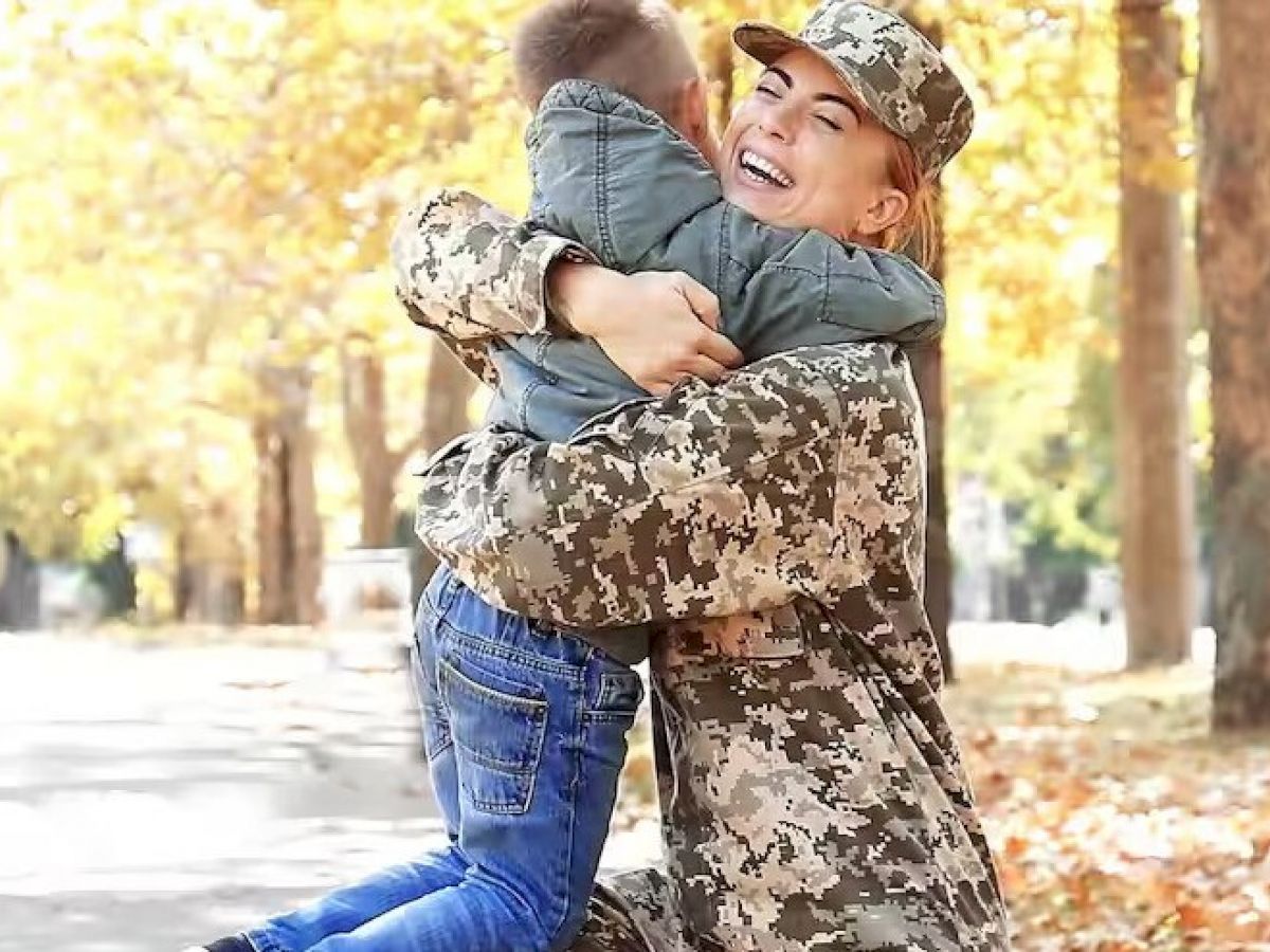 A woman in military uniform is joyfully hugging a young boy in a park, surrounded by autumn foliage and a sunny, tree-lined path.