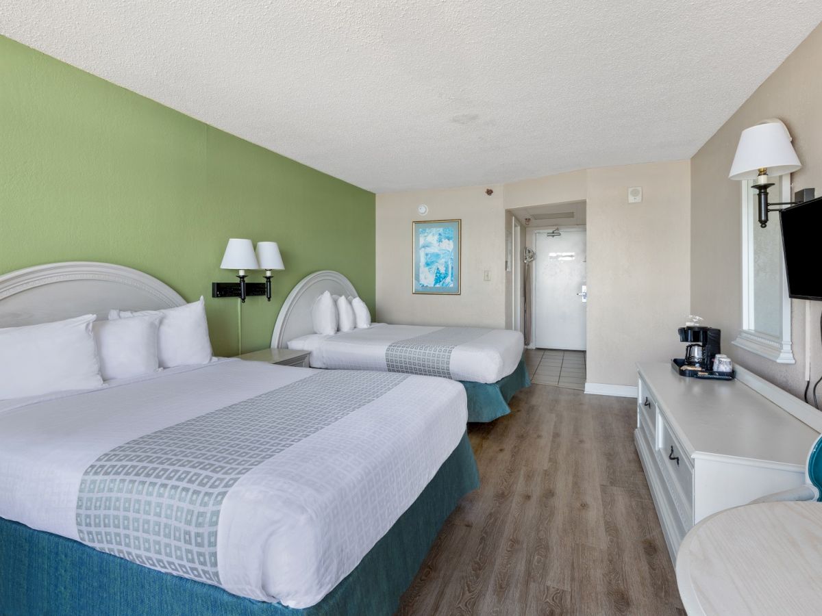 This image shows a hotel room with two queen-sized beds, a TV, coffee maker, wall art, and a small table with chairs, featuring wooden flooring.